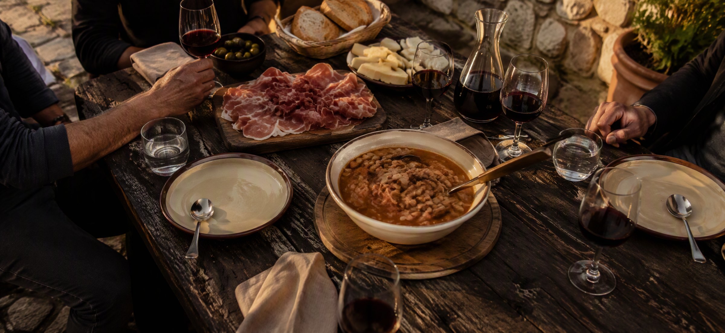 Rustic karst feast table in a stone courtyard with pršut, jota and glasses of Teran catching golden light
