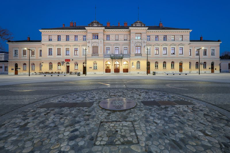 Piazza Transalpina / Trg Evrope at dusk, showing the illuminated historic building and the border marker