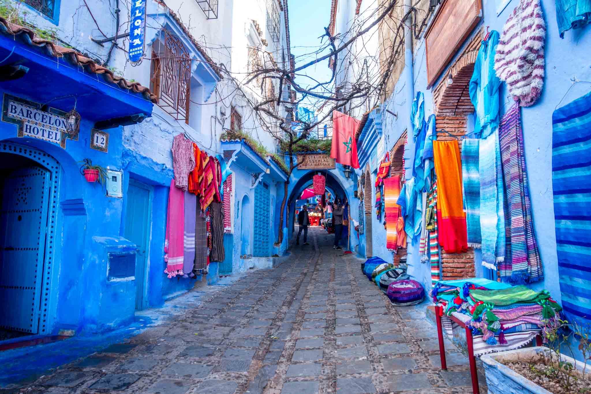 A cobbled street in Chefchaouen's medina lined with blue facades and colourful hanging textiles